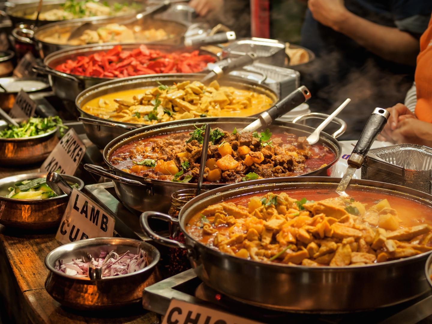 Various steaming Indian curries including lamb curry and chicken korma served in metal bowls at a food stall.