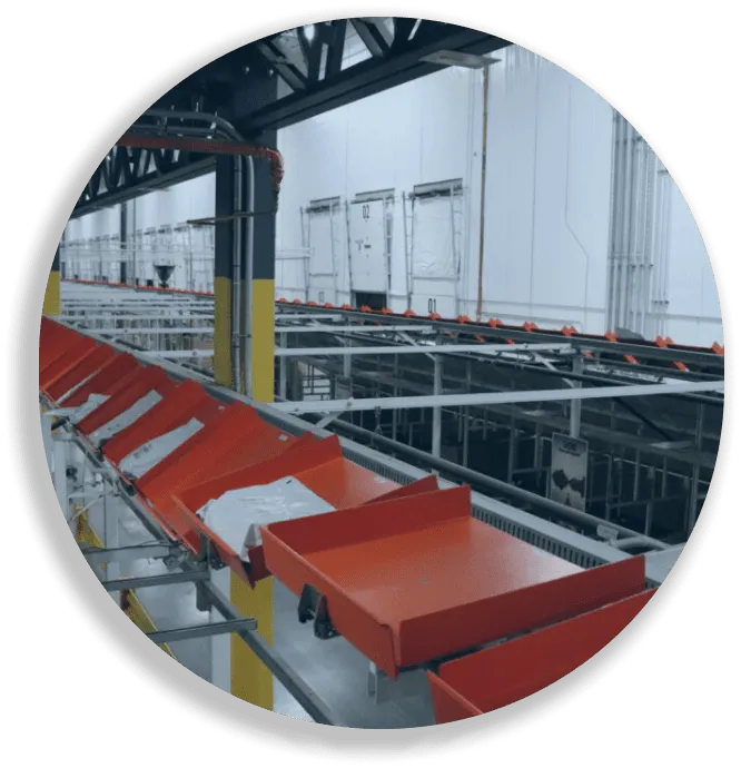 Industrial conveyor system with red bins in a large warehouse or factory setting.