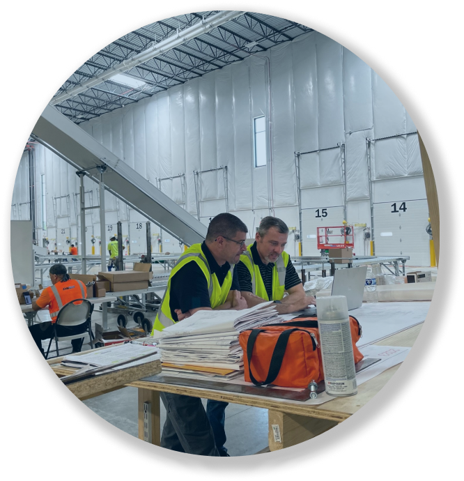 Two men in yellow vests review papers and a laptop inside a large warehouse with loading docks.