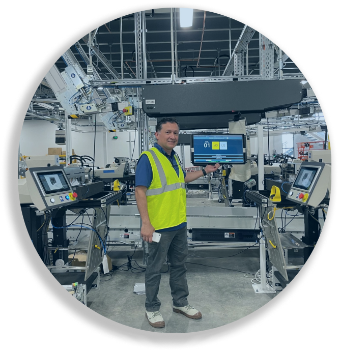 Man in yellow safety vest pointing at a large monitor in a high-tech manufacturing facility.