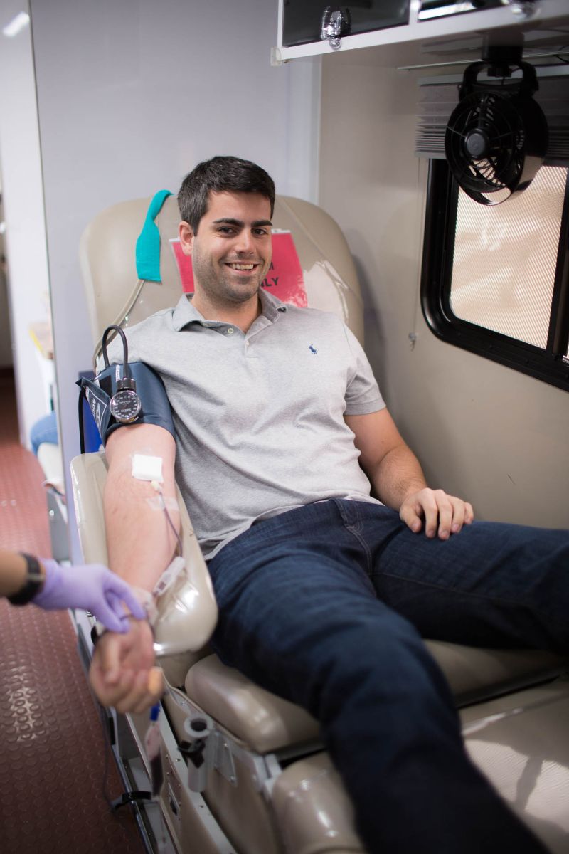 Smiling man donating blood seated on a medical chair with an IV in his arm and a pressure cuff.