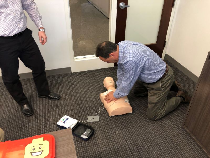 Man kneeling on carpeted floor performing CPR chest compressions on a training dummy in an office.