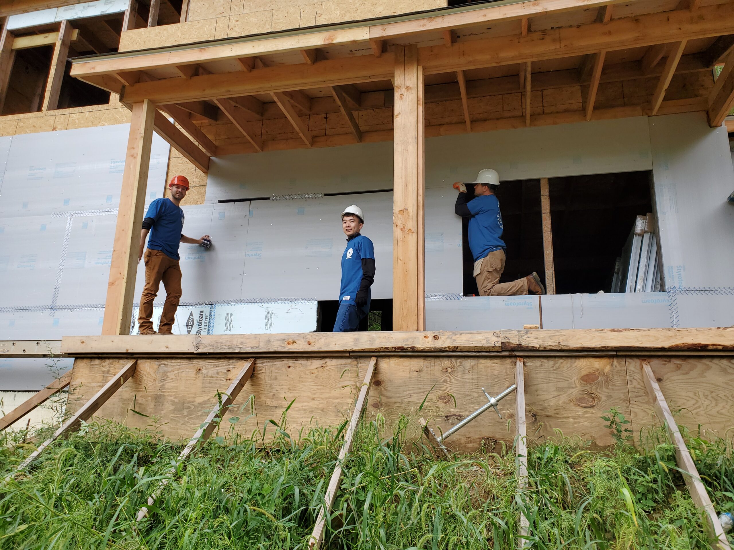 Three construction workers in blue shirts and helmets working on a wooden building frame.