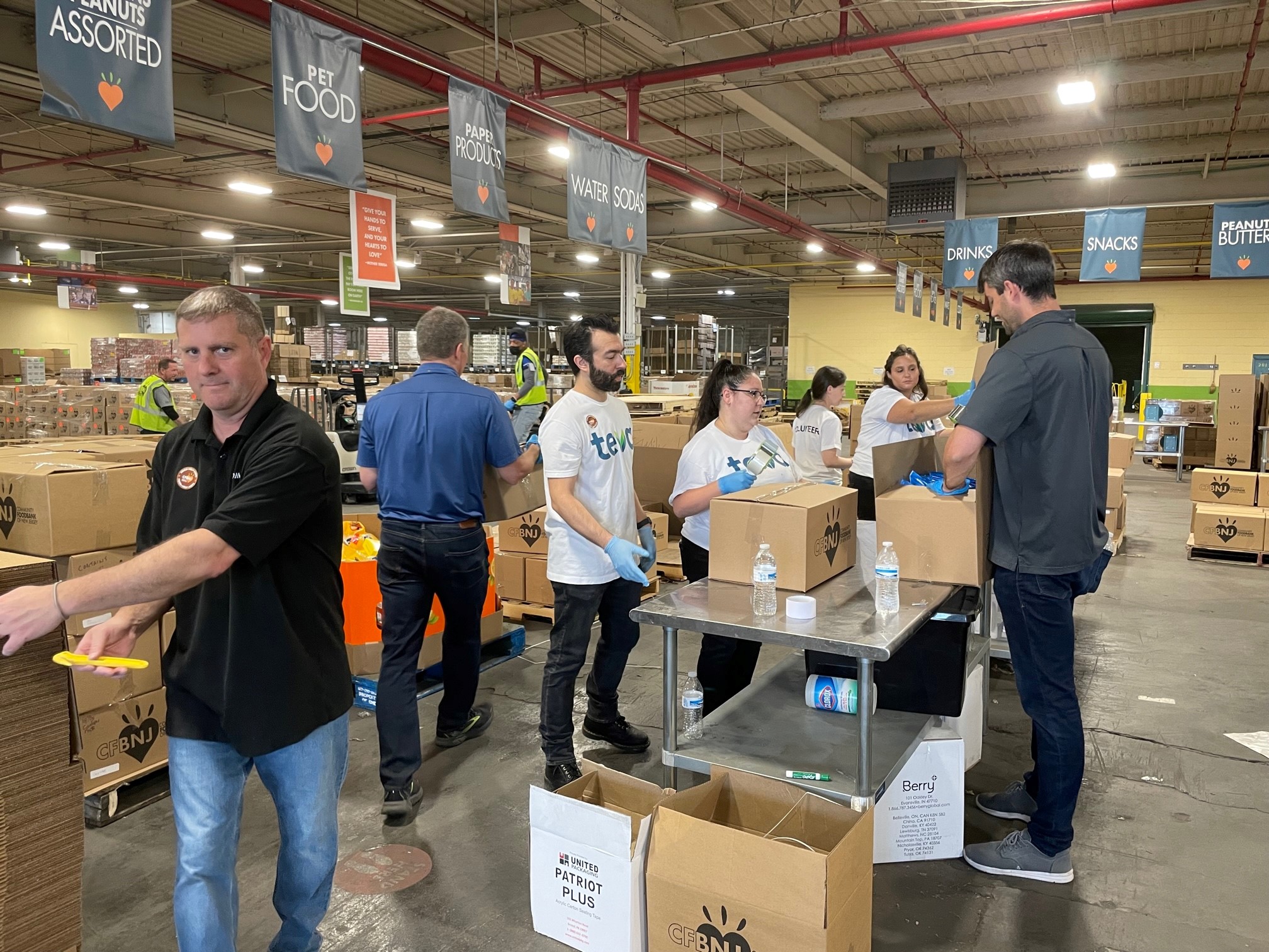 Volunteers packing food and supplies into boxes inside a large food bank warehouse.