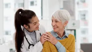 Female doctor comforting smiling elderly woman by gently holding her shoulder in a bright room.