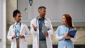 Three healthcare professionals in white coats and scrubs discussing in a hospital corridor.
