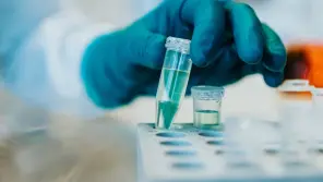 Gloved hand holding a test tube with green liquid in a laboratory setting.