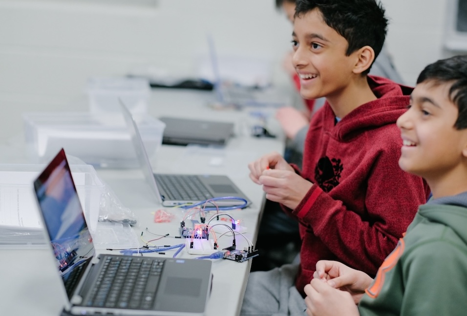 Two boys sit beside open laptops at a white classroom table, working with a small circuit board and tangled wires. The bright, softly blurred room and cool gray surfaces create a clean, focused STEM workshop scene.