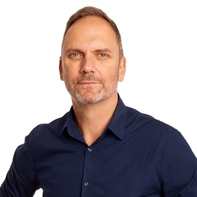 Portrait of a middle-aged man with short light brown hair and stubble wearing a dark blue buttoned shirt, looking directly at the camera with a neutral expression.
