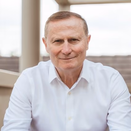 Portrait of a middle-aged man with light skin and short light brown hair, wearing a white button-up shirt, smiling slightly while seated outdoors with a blurred background.