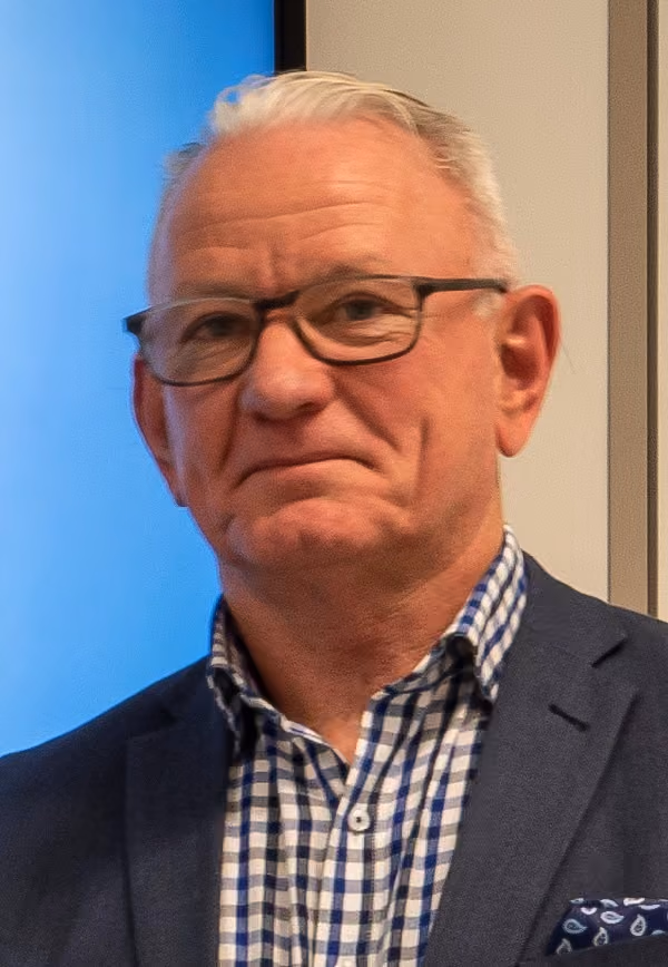Older man with short white hair and glasses wearing a checkered shirt and dark blazer, standing indoors against a neutral background with a blue section on the left.