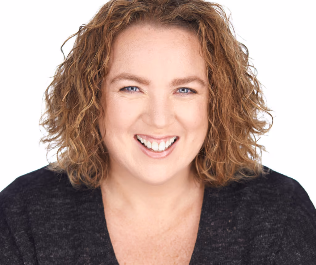 Smiling woman with curly light brown hair and blue eyes wearing a black top against a white background.