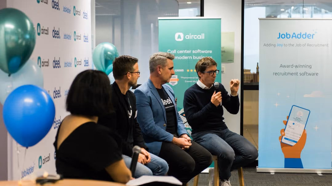 Four people in a panel discussion seated indoors with backdrop banners for Aircall and JobAdder, and blue and green balloons on the left side.