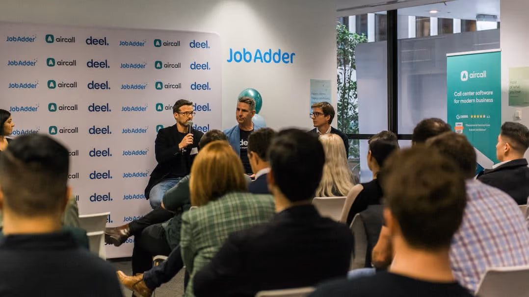 Three men seated at the front of a room are engaged in a panel discussion, one holding a microphone. The audience listens attentively. A backdrop displays logos of JobAdder, Aircall, and Deel. A green Aircall banner on the right promotes call center software for modern businesses.