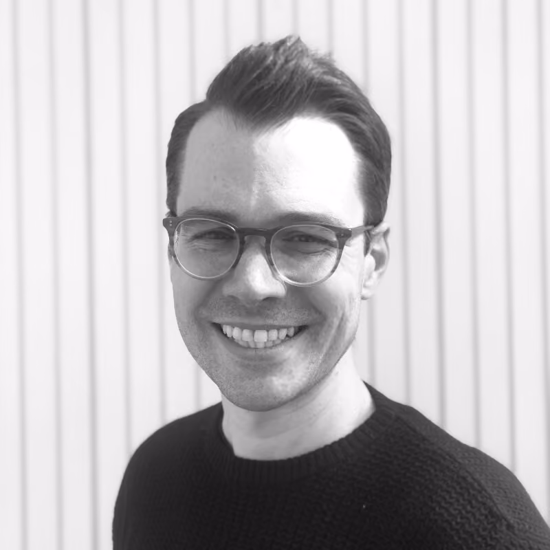 Smiling man with dark hair, glasses, and a black knit sweater standing in front of a vertical striped light background.