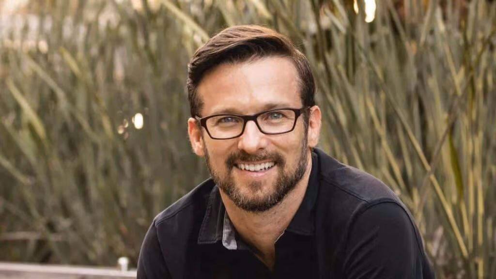 Smiling man with short brown hair, beard, and glasses wearing a black jacket poses outdoors in front of tall green grasses.