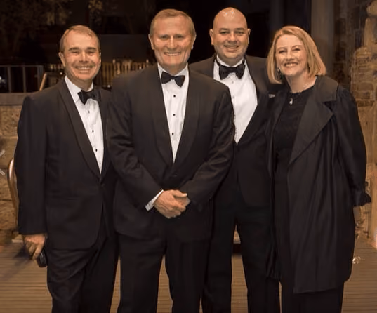 Four adults dressed formally in black tuxedos and a black dress with coat, standing indoors and smiling at the camera.