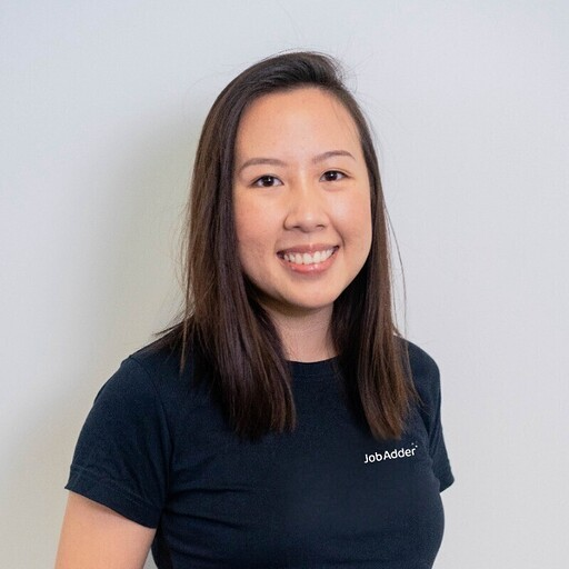 Smiling young woman with straight shoulder-length dark hair wearing a black JobAdder t-shirt standing against a plain light gray background.