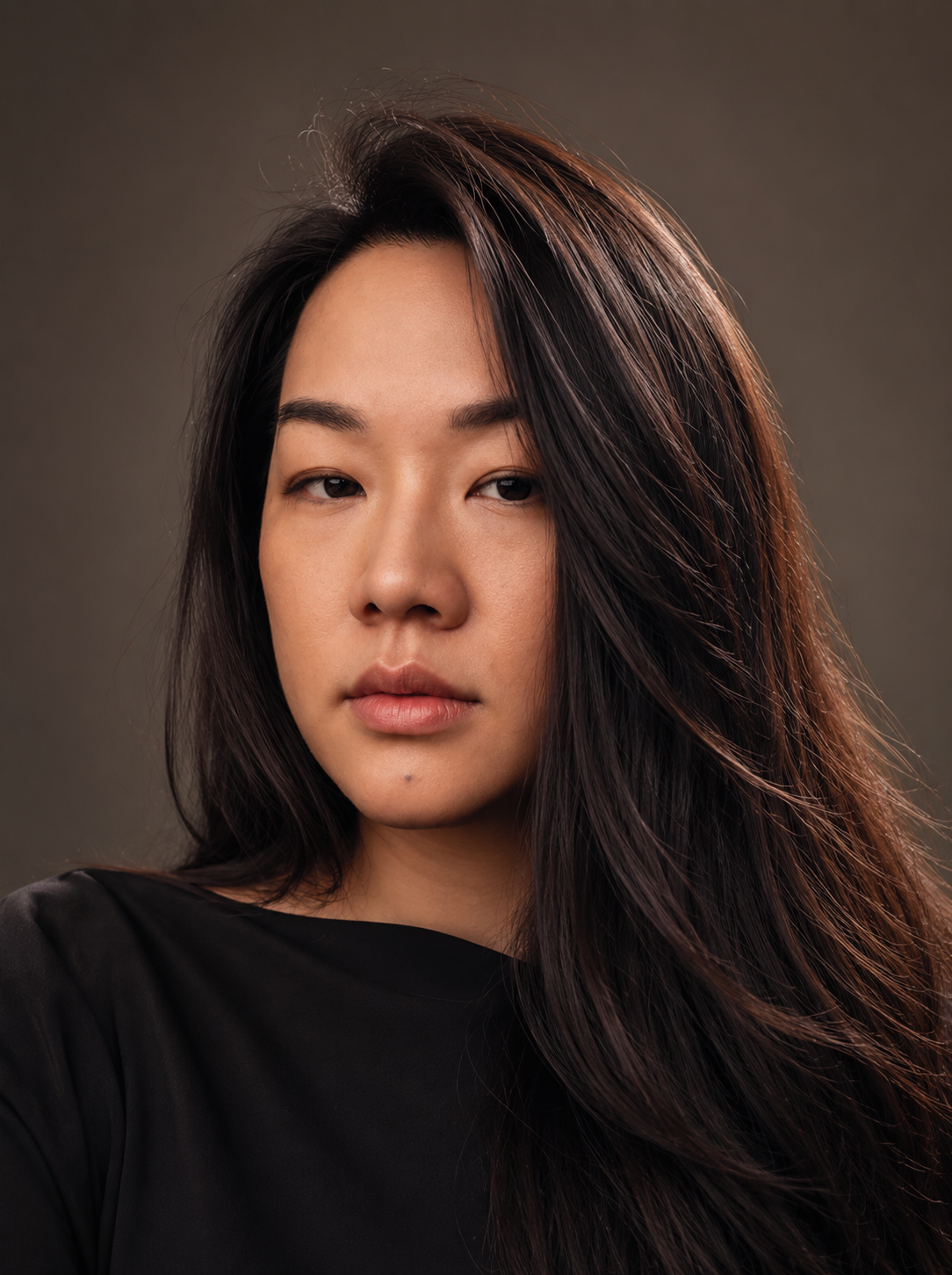 Close-up portrait of a young woman with long dark hair wearing a black top against a neutral brown background, looking calmly at the camera.