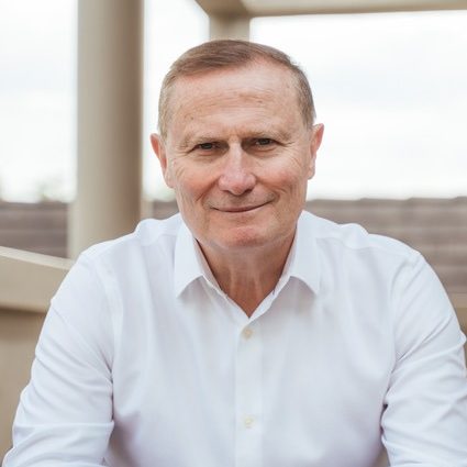 Middle-aged man with short light brown hair wearing a white button-up shirt, looking at the camera with a slight smile, seated outdoors with a blurred background.