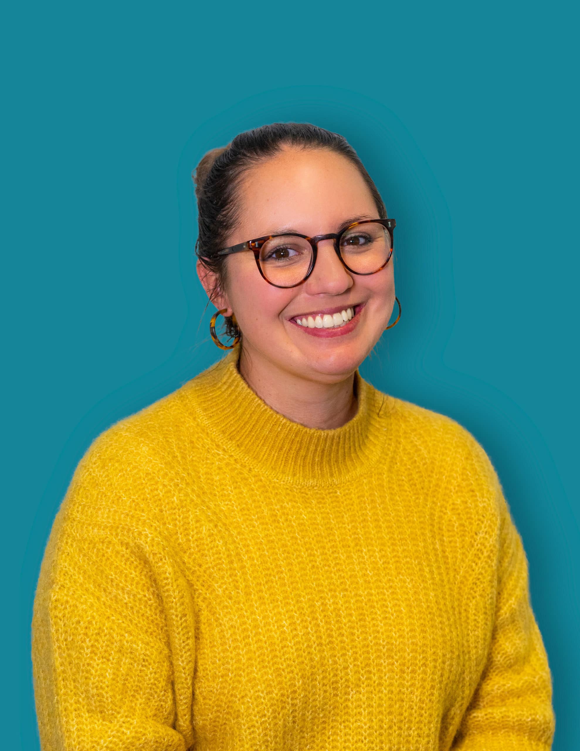 Smiling woman with dark hair tied back, wearing tortoiseshell glasses and hoop earrings, dressed in a yellow knitted sweater, with a teal background.