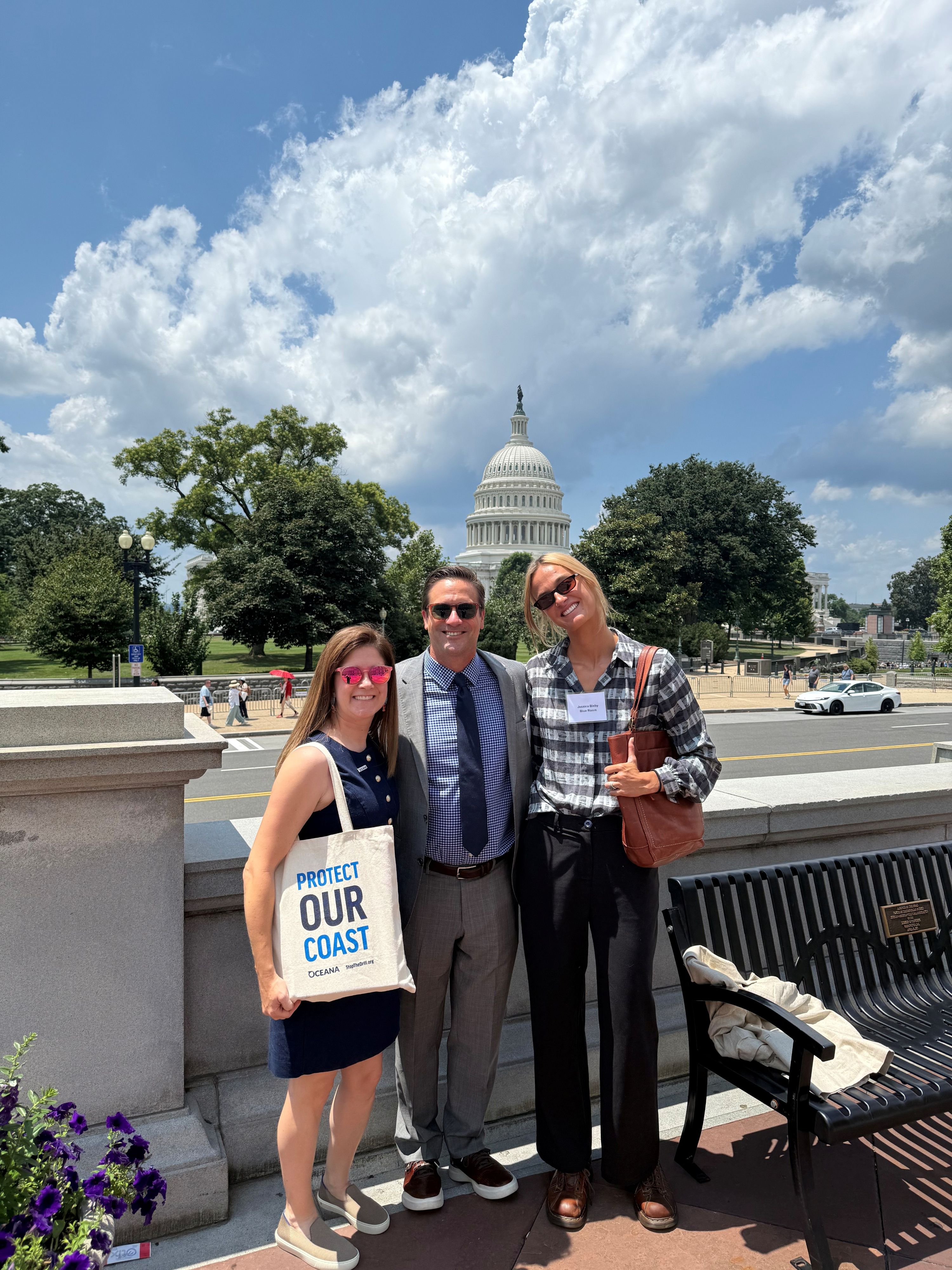 Three people smiling in front of a State Capitol building with a clear sky and white clouds above.
