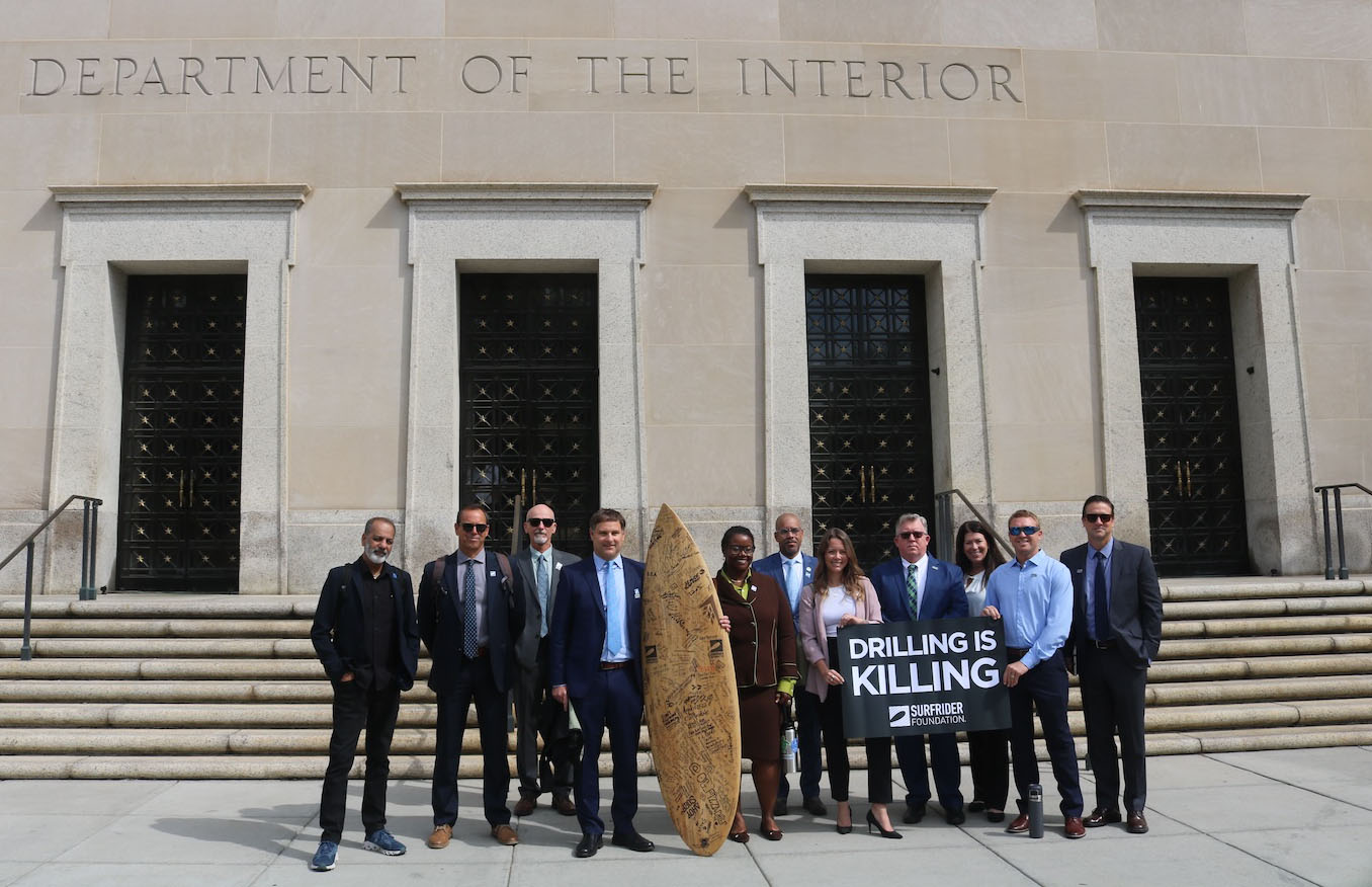 A group of professionally dressed people standing in front of the Department of The Interior building holding a surfboard and a sign that reads "Drilling is Killing"