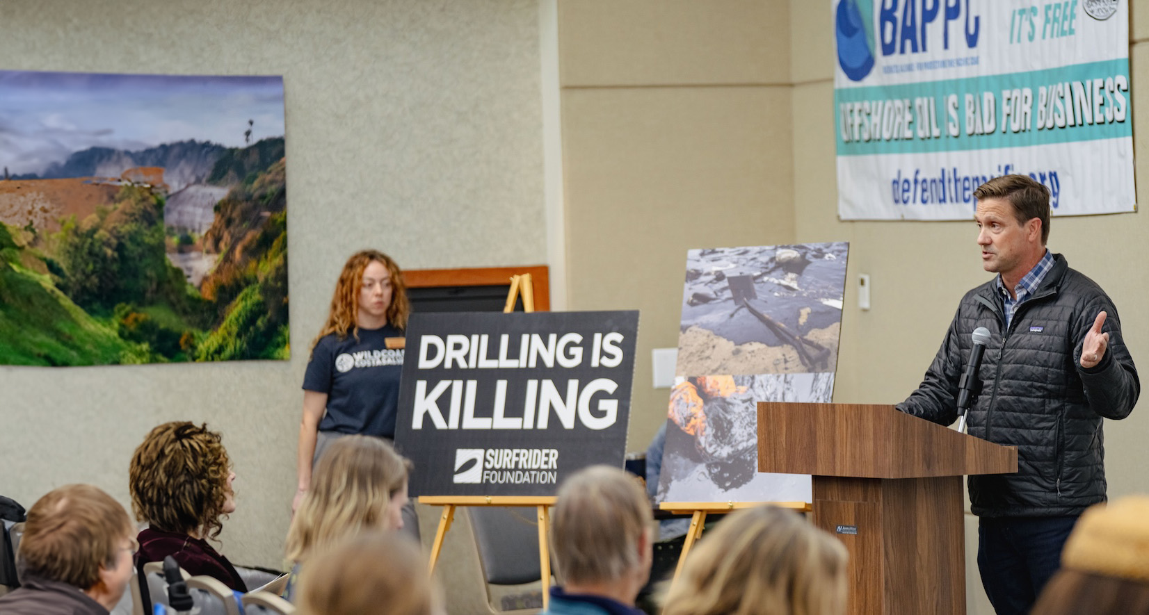 Man speaking at a podium during a protest meeting with signs against offshore oil drilling.