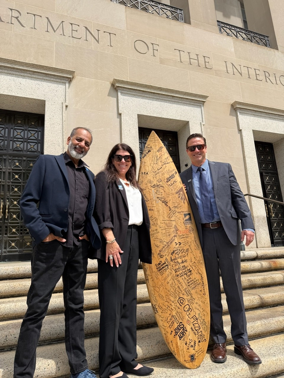 Three professionally dressed people standing on steps holding a surfboard with signatures written on it in front of a building.