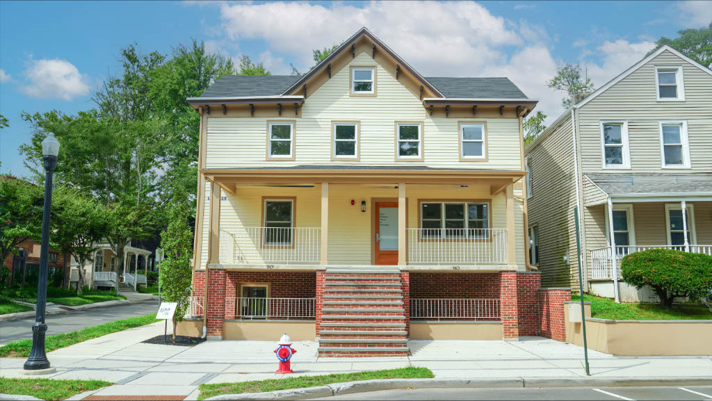 Front view of a two-story yellow house with a brick porch base, orange door, and central steps, set behind a sidewalk with a fire hydrant.