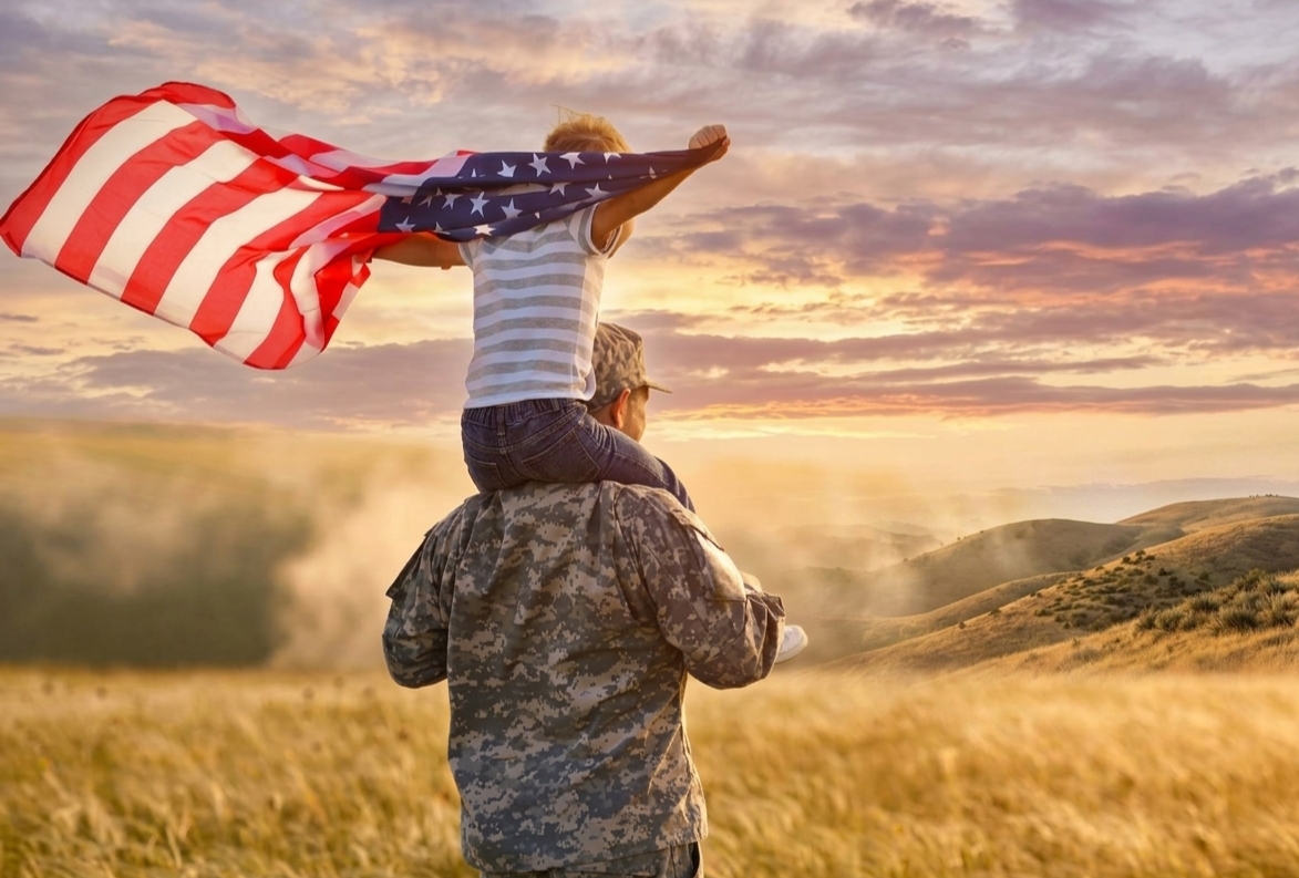 military service member walking with child on his shoulders and American flag waving