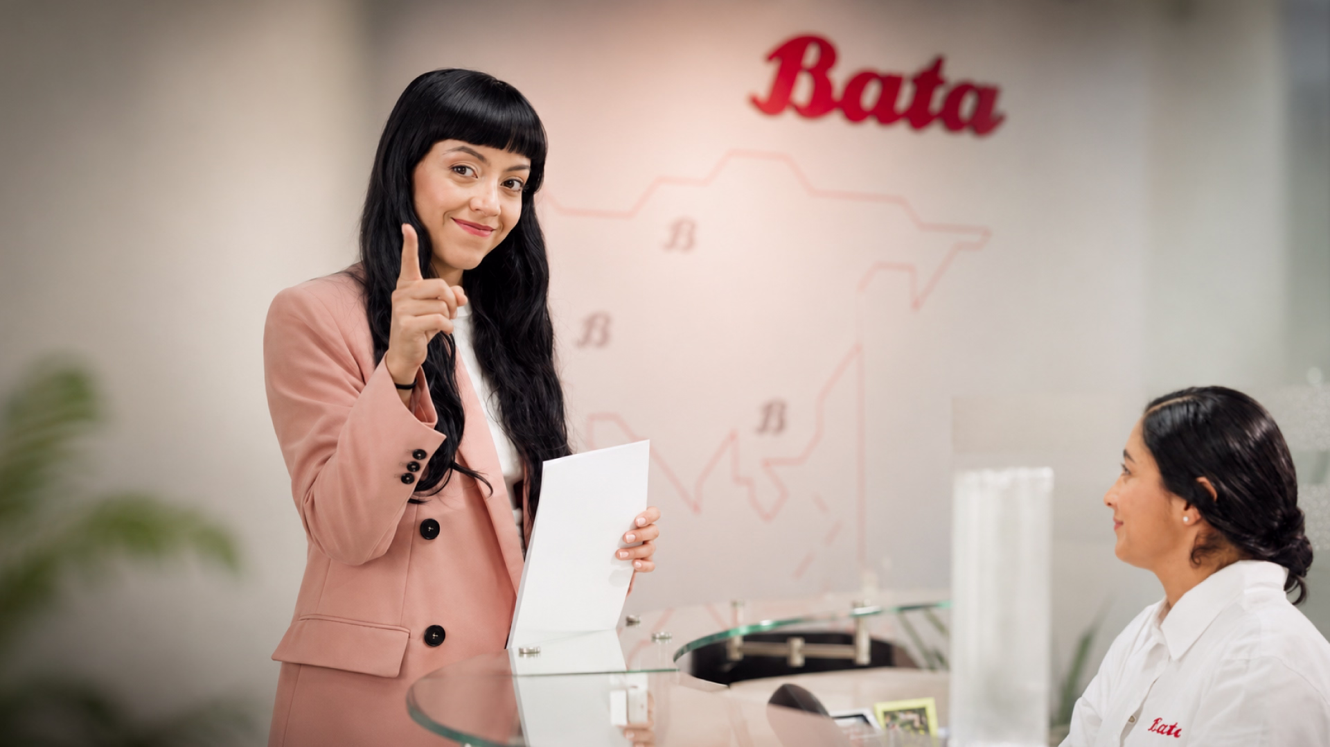 Mujer de saco rosa señalando a cámara con documentos en mano en recepción de oficinas Bata, junto a recepcionista sonriente