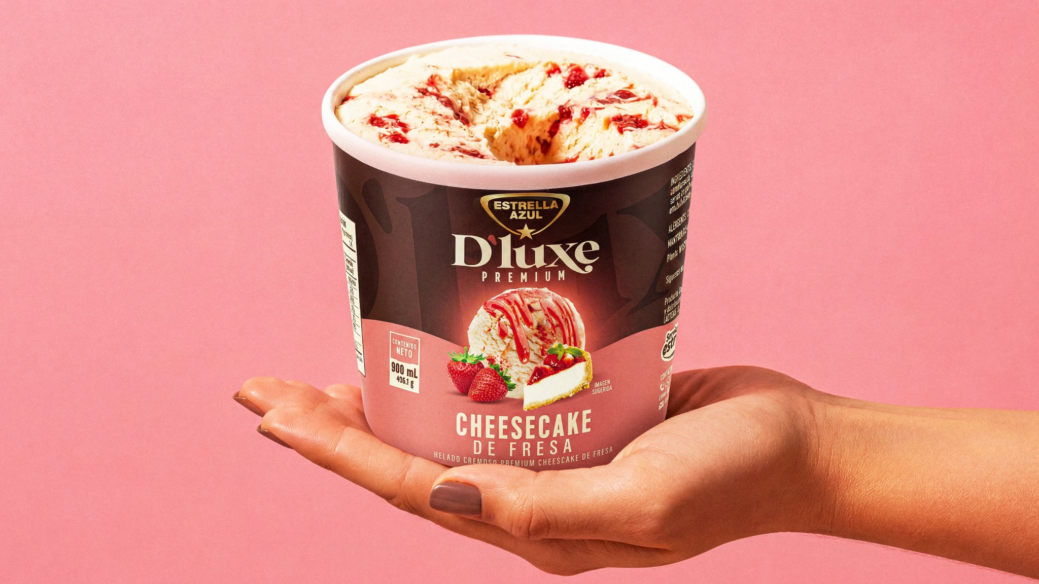 Mano sosteniendo un envase abierto de helado premium D'luxe Cheesecake de Fresa de Estrella Azul sobre fondo rosa.