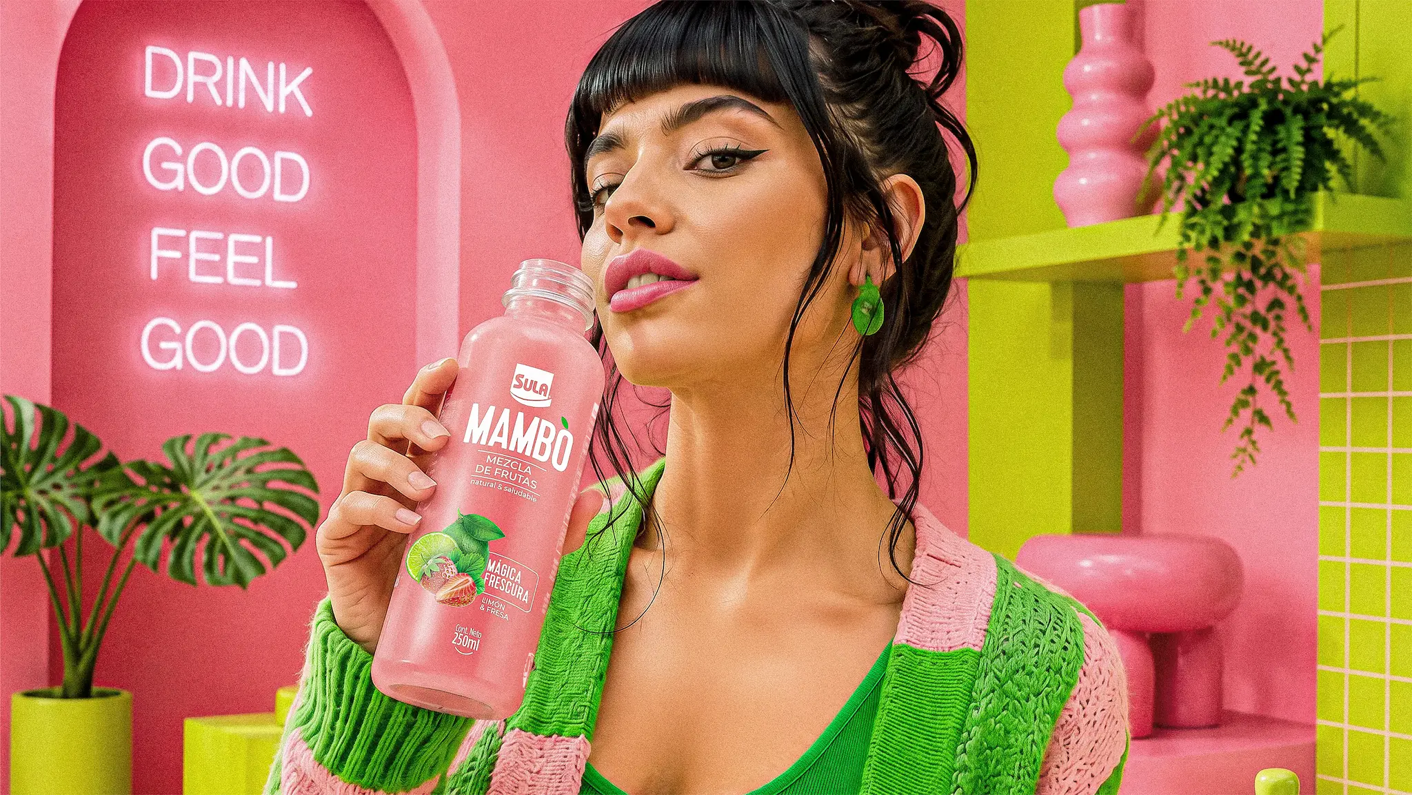 Mujer sosteniendo una botella Mambo Mágica Frescura frente a letrero de neón Drink Good Feel Good en escenario rosa y verde