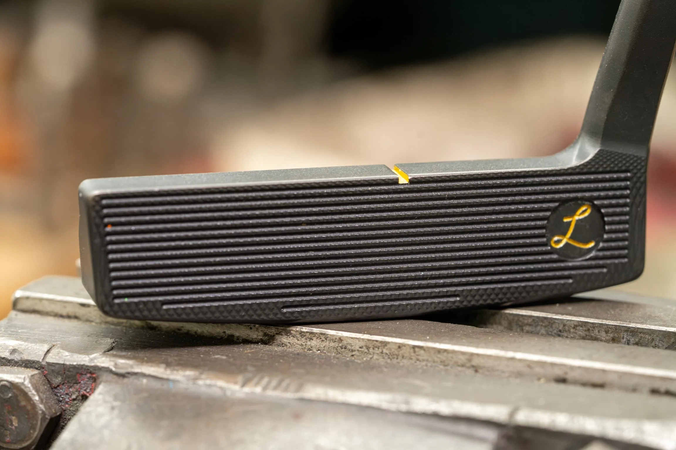 Close-up of a black Lajosi putter face with precision-milled grooves, a gold alignment mark and maker emblem, highlighting the detailed workshop finish behind each custom club.