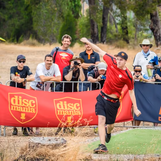 Disc golfer drives from the tee as spectators and photographers watch behind Discmania barriers, capturing the competitive energy, audience interest and professional event atmosphere of the Aussie Open.