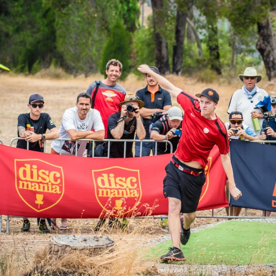 Disc golfer drives from the tee as spectators and photographers watch behind Discmania barriers, capturing the competitive energy, audience interest and professional event atmosphere of the Aussie Open.