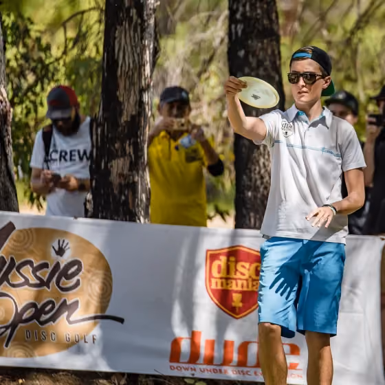 Disc golfer drives from the tee as spectators and photographers watch behind Discmania barriers, capturing the competitive energy, audience interest and professional event atmosphere of the Aussie Open.
