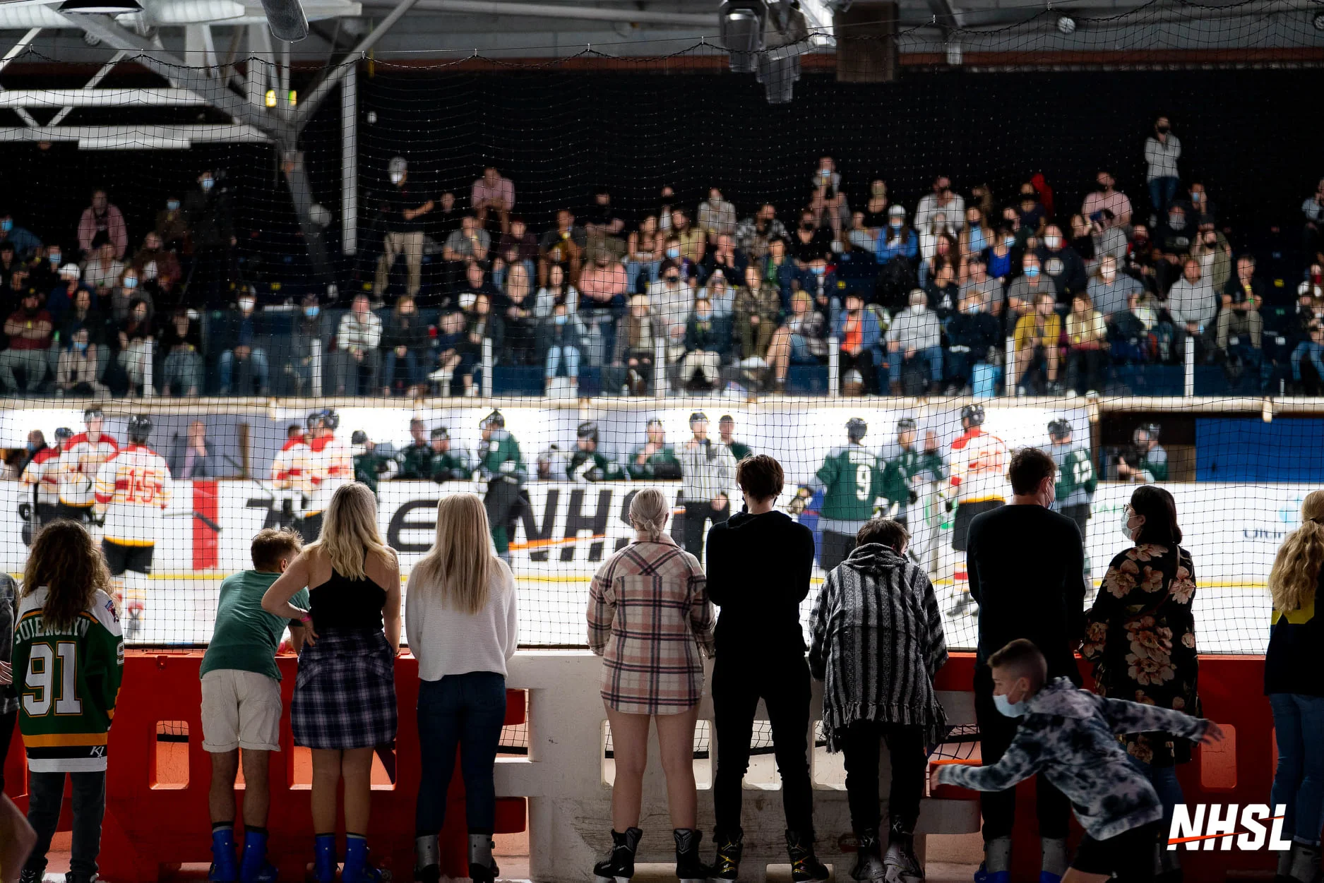 Fans watch an NHSL game from rinkside and packed stands as players gather on the ice, showing the strong attendance, shared atmosphere and fan experience created around the new national league.