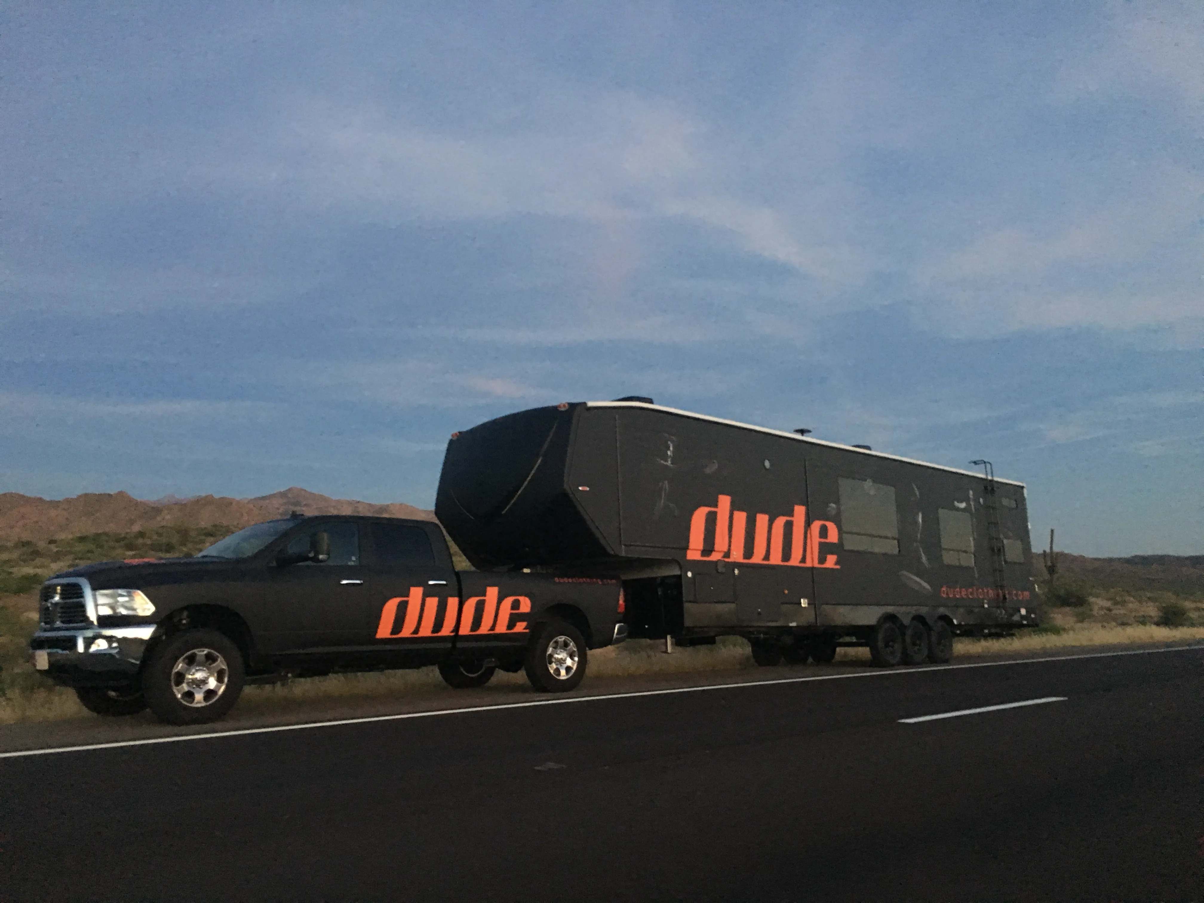 DUDE-branded truck and large touring trailer parked beside an open highway, showing the apparel brand’s bold identity and hands-on presence within the disc golf community.