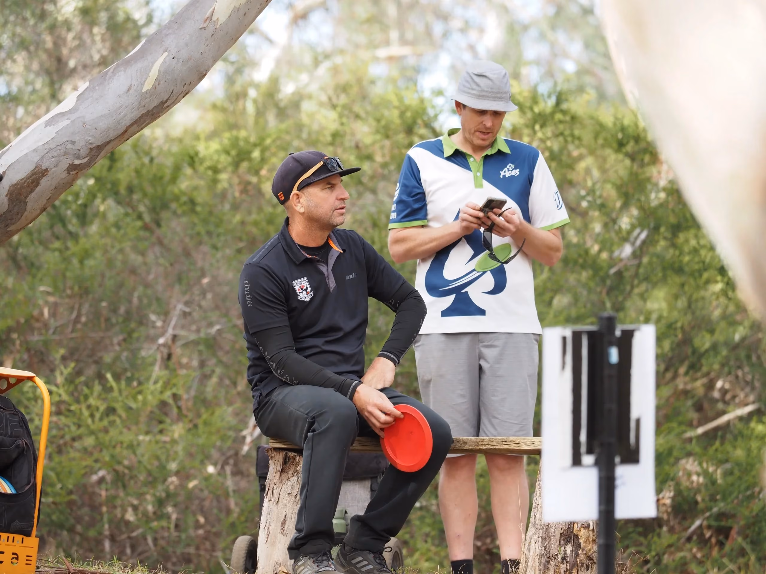 Team Aces disc golfer waits with a red disc while a teammate checks scores on a phone, showing the mix of competition, coordination and community behind the brand’s social content.