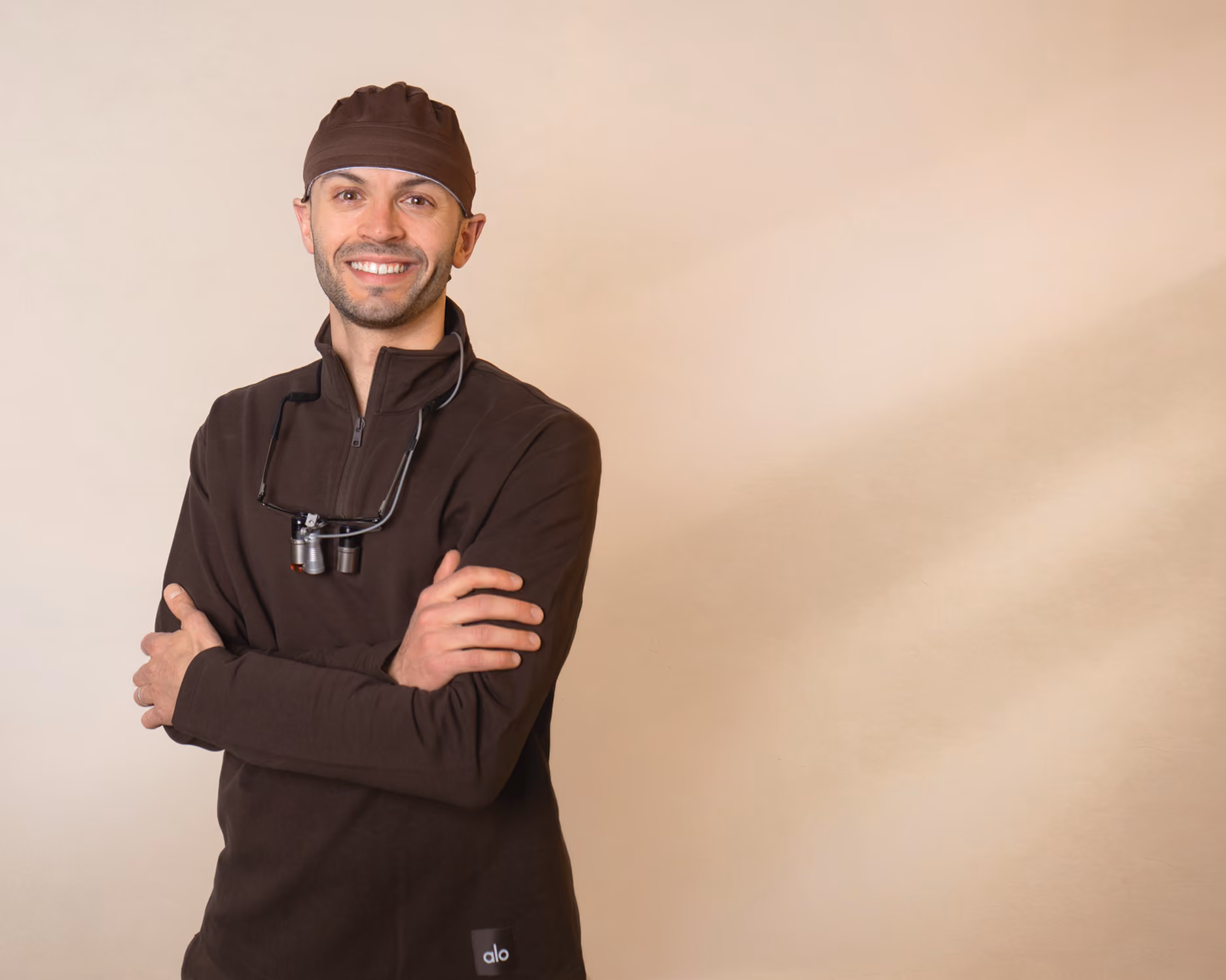Smiling man wearing a dark cap and pullover with magnifying glasses around his neck, arms crossed.