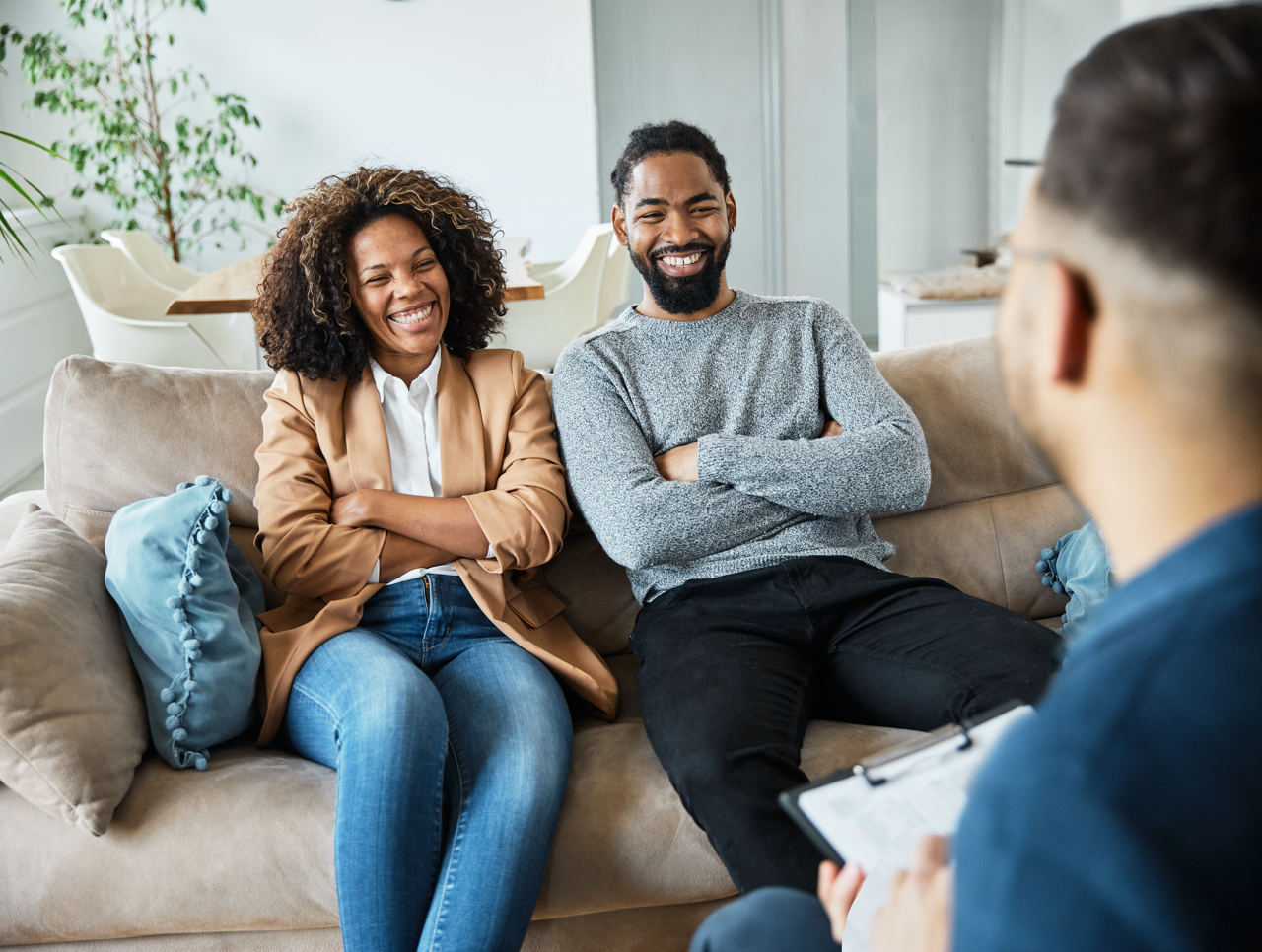 Smiling couple sitting on a beige couch with arms crossed, engaged in conversation with a person facing them holding a clipboard in a bright room.