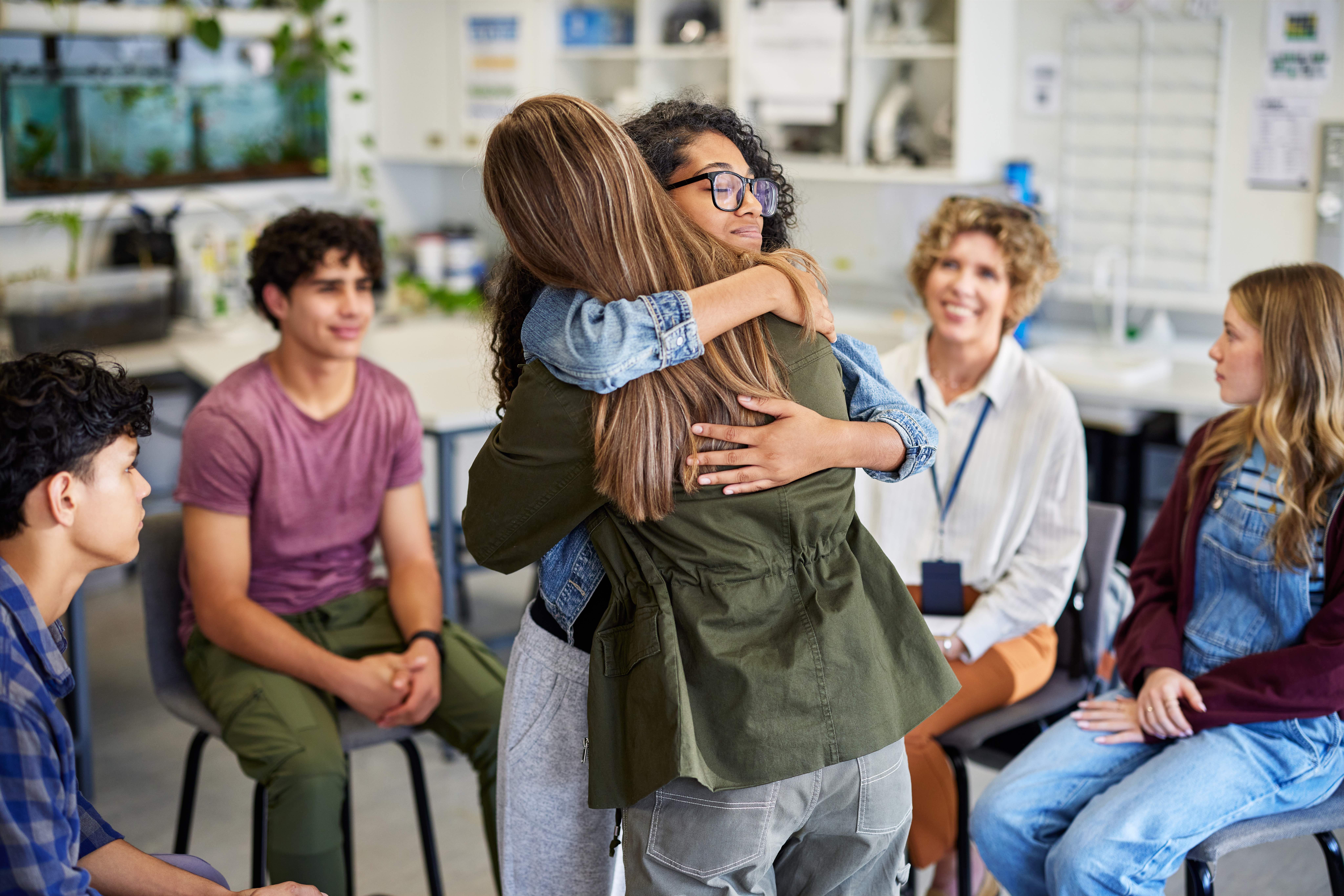 Two young women warmly hugging in a supportive group meeting, surrounded by smiling and attentive peers seated in a circle in a bright, informal room.