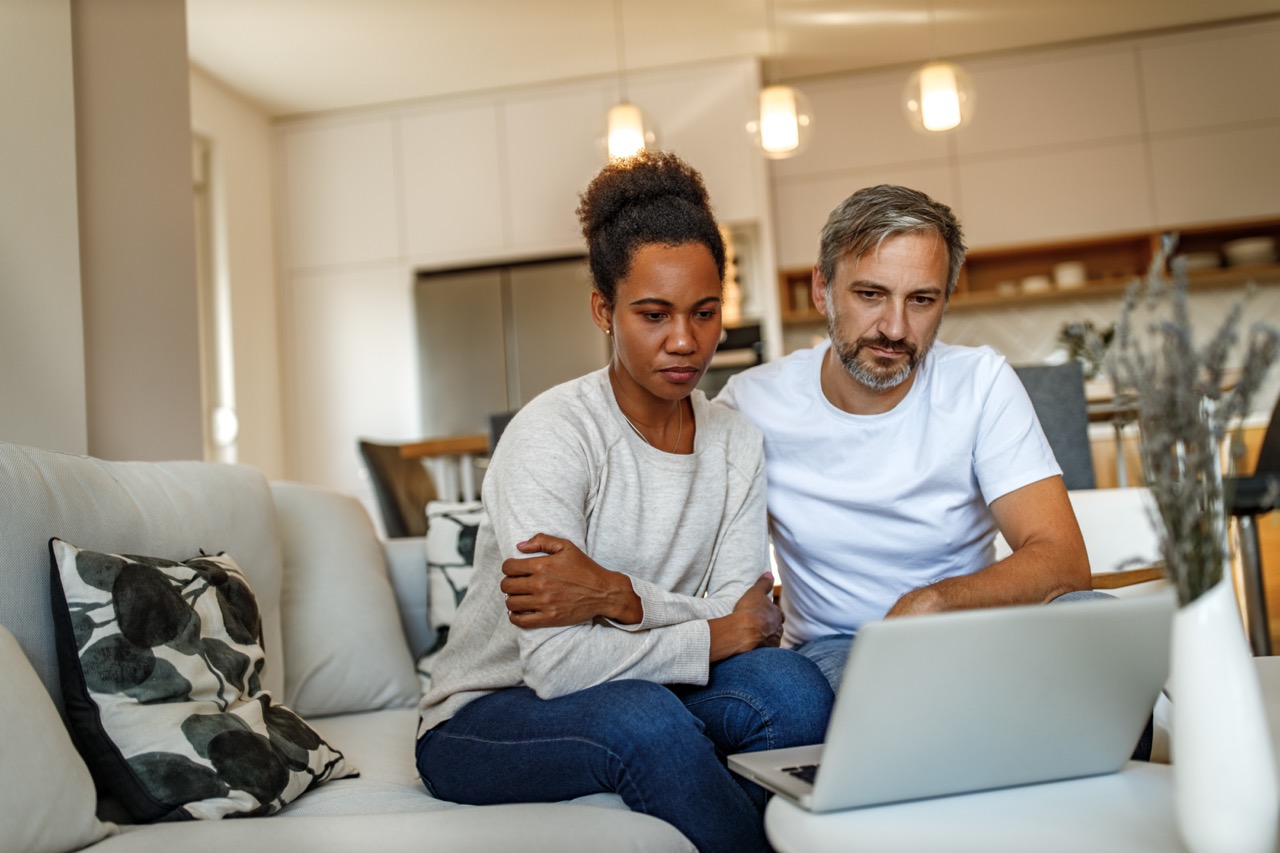 A couple sitting closely on a light-colored couch in a modern living room, focused on a laptop on a small table in front of them.