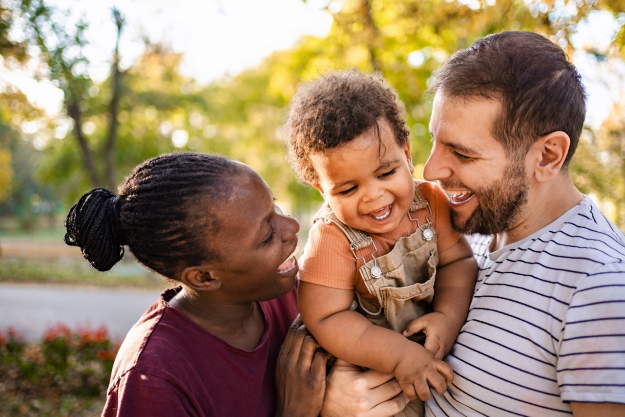 Smiling mixed-race family outdoors with a woman holding a laughing toddler and a man in a striped shirt closely engaged, surrounded by green trees.