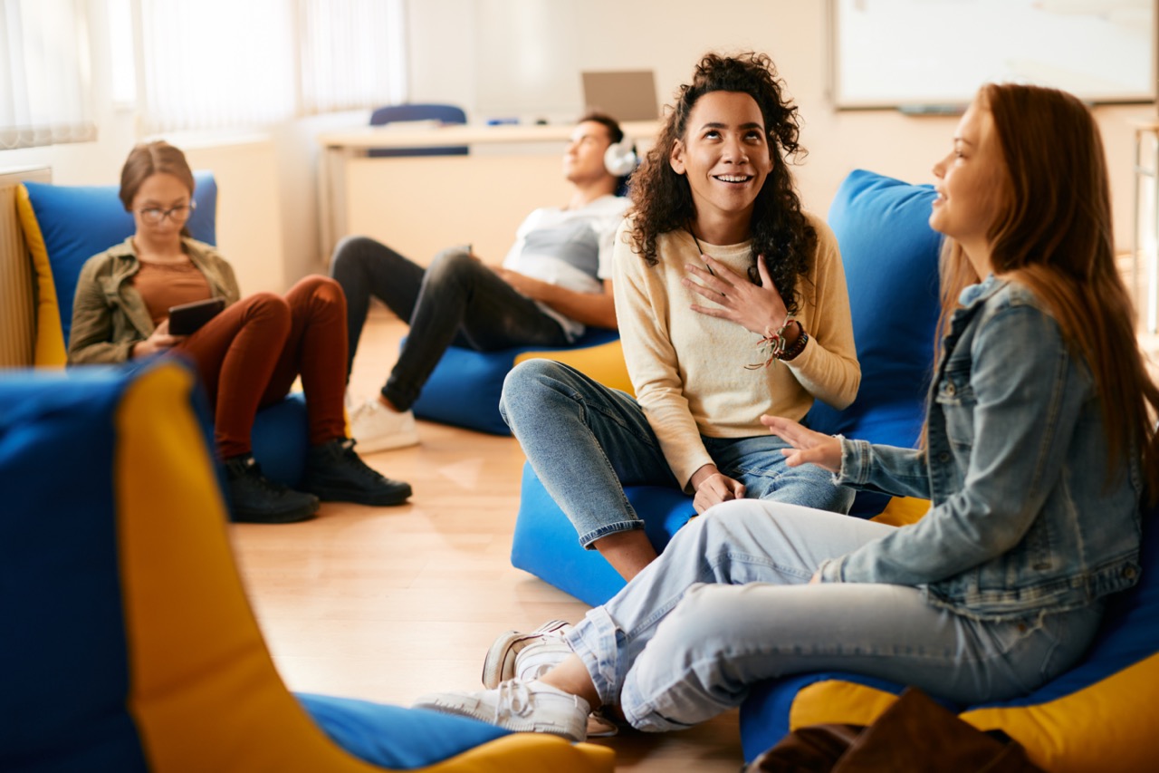 Four young adults sitting on blue and yellow bean bags in a casual, well-lit workspace; two women in the foreground are talking and smiling, while a woman with glasses reads a tablet and a man with headphones relaxes in the background.