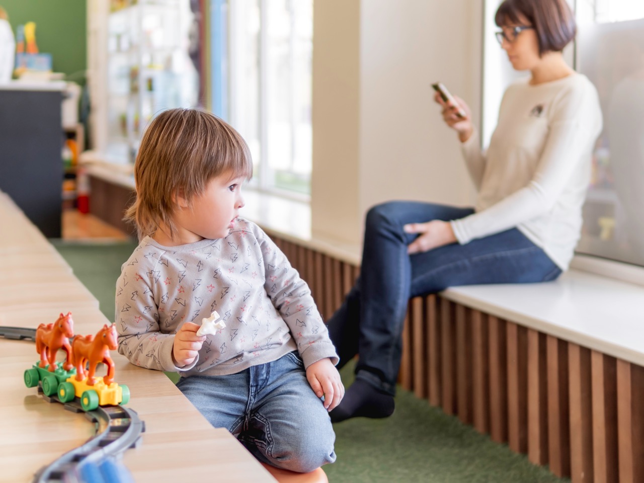 A toddler kneeling on a bench playing with a toy train set with horse figurines, while an adult sitting on a nearby window seat looks at a smartphone.