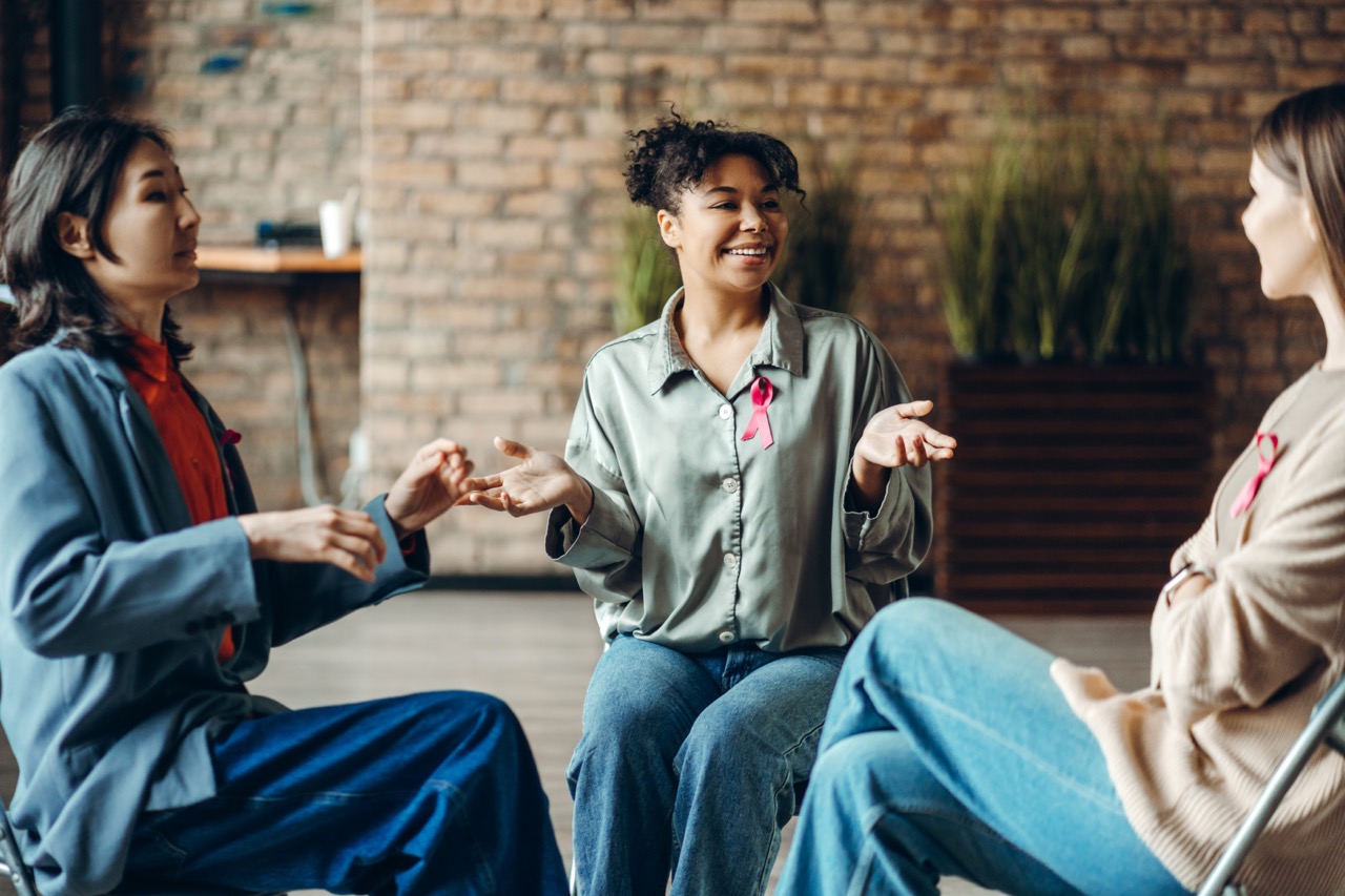 Three diverse women sitting in a circle indoors, engaged in conversation and smiling, each wearing a pink breast cancer awareness ribbon.
