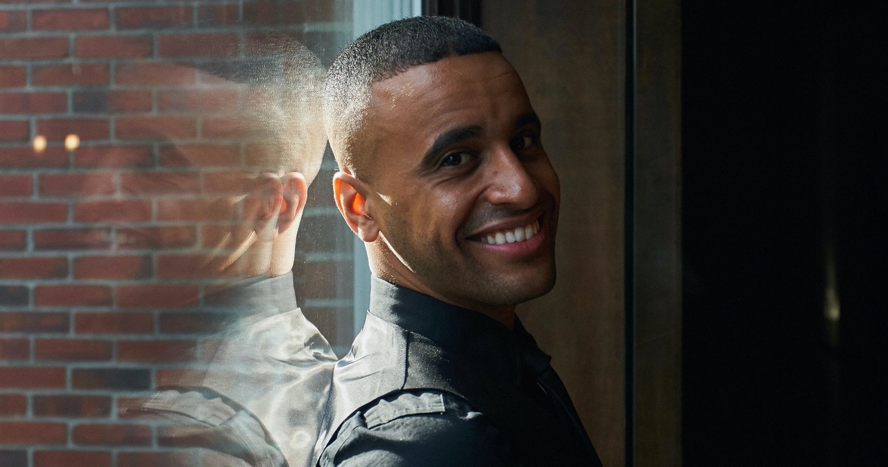 Smiling man with short hair wearing a black shirt, standing near a window with his reflection visible on the glass.